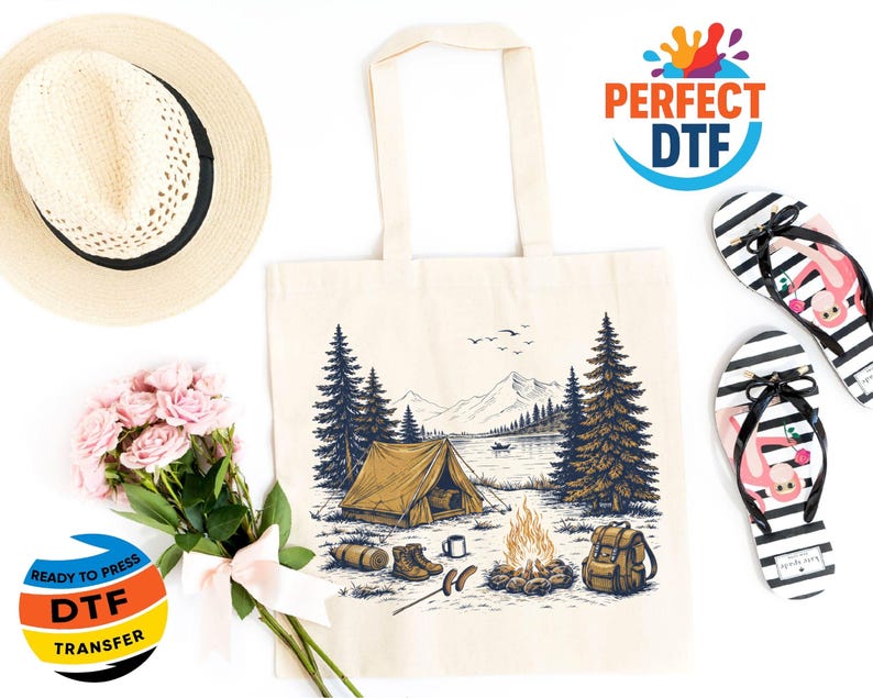May include: Cream-colored tote bag with a camping scene illustration, featuring a tent, campfire, and mountains. The image includes a straw hat, pink flowers, and black and white striped flip-flops with a flamingo design. The text "READY TO PRESS DTF TRANSFER" is visible.