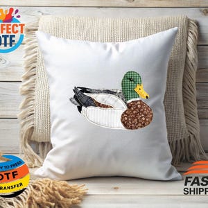 May include: White square pillow with a detailed appliqu&eacute; of a duck. The duck features a green and white checkered head, a brown floral patterned body, and a mix of gray, white, and black feathers. The pillow is on a beige fringed cushion.
