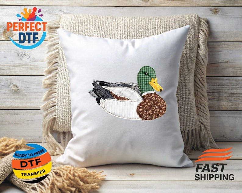 May include: White square pillow with a detailed appliqu&eacute; of a duck. The duck features a green and white checkered head, a brown floral patterned body, and a mix of gray, white, and black feathers. The pillow is on a beige fringed cushion.