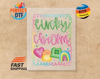 May include: A colorful graphic with the words "Lucky Charm" in green and pink script. The design includes a pink heart, a pot of gold, a green shamrock, and a rainbow. The image is a ready-to-press DTF transfer.