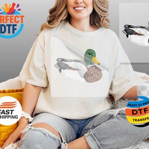 May include: Cream-colored t-shirt featuring a detailed duck design. The duck has a green plaid head, a brown patterned body, and gray and white wings. The design is on the front of the shirt.