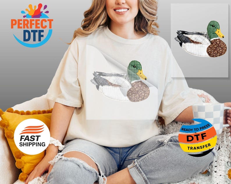 May include: Cream-colored t-shirt featuring a detailed duck design. The duck has a green plaid head, a brown patterned body, and gray and white wings. The design is on the front of the shirt.