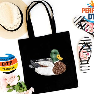 May include: Black tote bag with a duck applique. The duck features green plaid, brown floral, and white fabric. Striped flip-flops with flamingo graphics and a straw hat are also visible.