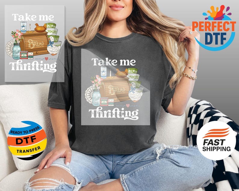 May include: Dark grey t-shirt with the text "Take me Thrifting" and a graphic of a bread box, bowls, flowers, and a duck. The shirt is worn by a person sitting on a white couch.