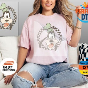 May include: A light pink t-shirt featuring a cartoon image of Goofy inside a checkered circle. The cartoon Goofy has a green hat and his tongue is sticking out. The shirt is worn by a person.