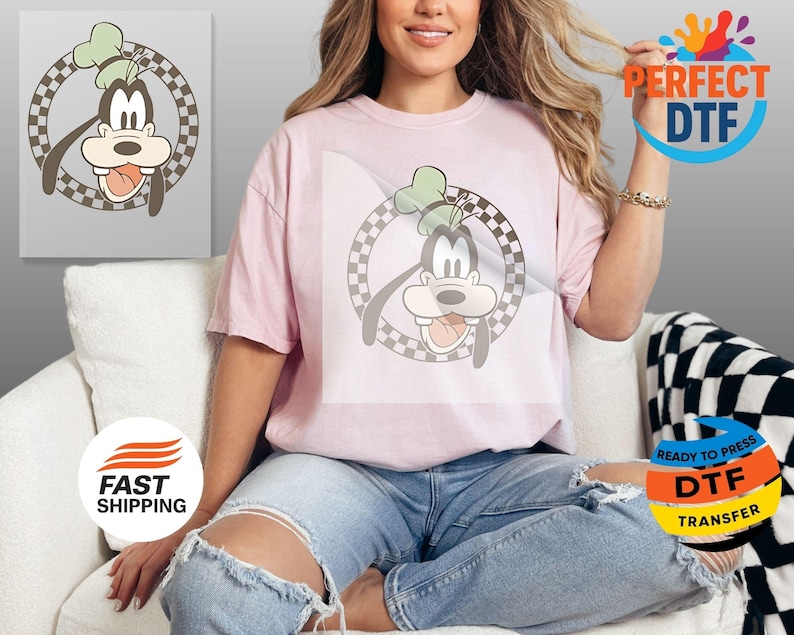May include: A light pink t-shirt featuring a cartoon image of Goofy inside a checkered circle. The cartoon Goofy has a green hat and his tongue is sticking out. The shirt is worn by a person.