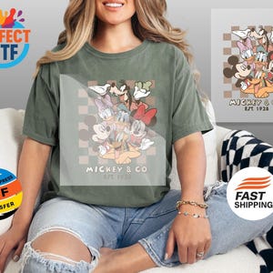 May include: An olive green t-shirt featuring a graphic of Mickey Mouse and friends, with the text "MICKEY & CO. EST 1928". The design includes Mickey, Minnie, Donald, Daisy, Goofy, and Pluto. The shirt is paired with distressed blue jeans.