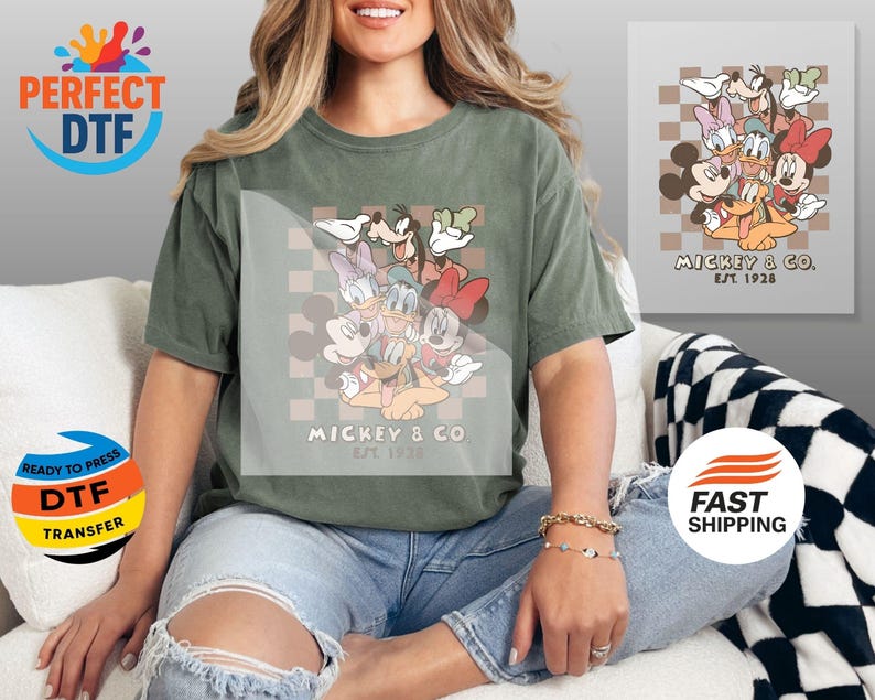 May include: An olive green t-shirt featuring a graphic of Mickey Mouse and friends, with the text "MICKEY & CO. EST 1928". The design includes Mickey, Minnie, Donald, Daisy, Goofy, and Pluto. The shirt is paired with distressed blue jeans.