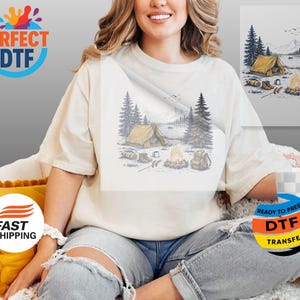 May include: Cream-colored t-shirt featuring a camping scene with a tent, campfire, and mountains. The graphic is in shades of blue, yellow, and brown. The shirt has a relaxed fit and is worn by a person.