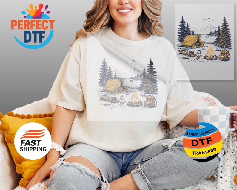 May include: Cream-colored t-shirt featuring a camping scene with a tent, campfire, and mountains. The graphic is in shades of blue, yellow, and brown. The shirt has a relaxed fit and is worn by a person.
