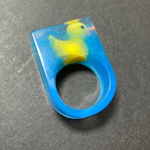 May include: A blue resin ring with a yellow rubber ducky embedded inside. The ducky is facing to the right and has a small beak and black eyes.