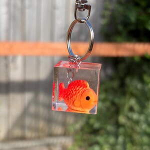 May include: A clear resin cube keychain featuring a small, bright orange fish figurine suspended inside. The cube is attached to a silver-toned metal split ring and a lobster claw clasp, creating a functional accessory with a transparent, glossy finish.