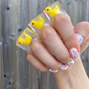 Könnte beinhalten: Drei klare Acrylringe, jeder mit einer gelben Quietscheente mit orangefarbenem Schnabel. Die Ringe werden hochgehalten, um das Design zu präsentieren. Der Hintergrund ist ein Holzzaun.