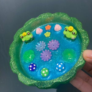 Pode incluir: Um prato decorativo de resina com uma cena de lagoa. O prato tem uma borda verde brilhante e um interior azul com figuras de sapos, flores, nenúfares e detalhes de bolinhas. O prato foi projetado para guardar itens pequenos.