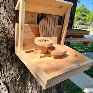 May include: A wooden bird feeder shaped like a miniature house with a small chair and table. The feeder is attached to a tree trunk and has a roof over the chair and table. There are peanuts on the table.