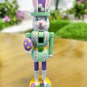 May include: A wooden Easter bunny nutcracker with a purple hat, green and white polka dot vest, and a purple and yellow egg in its hand. The nutcracker is standing on a yellow base with three colorful eggs and a small bunny figurine.