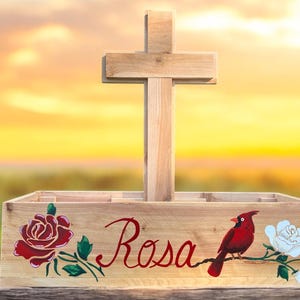 May include: Wooden cross-shaped memorial planter with the name "Rosa" in cursive. The planter features painted red roses, a white rose, and a red cardinal bird. The background is a blurred sunset.