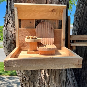 May include: A wooden bird feeder with a small wooden chair and table attached to it. The feeder is mounted on a tree trunk. There are a few nuts on the table.