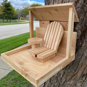 May include: A wooden bird feeder with a small chair attached to the front. The feeder has a roof and a tray for birdseed. The feeder is mounted to a tree trunk.