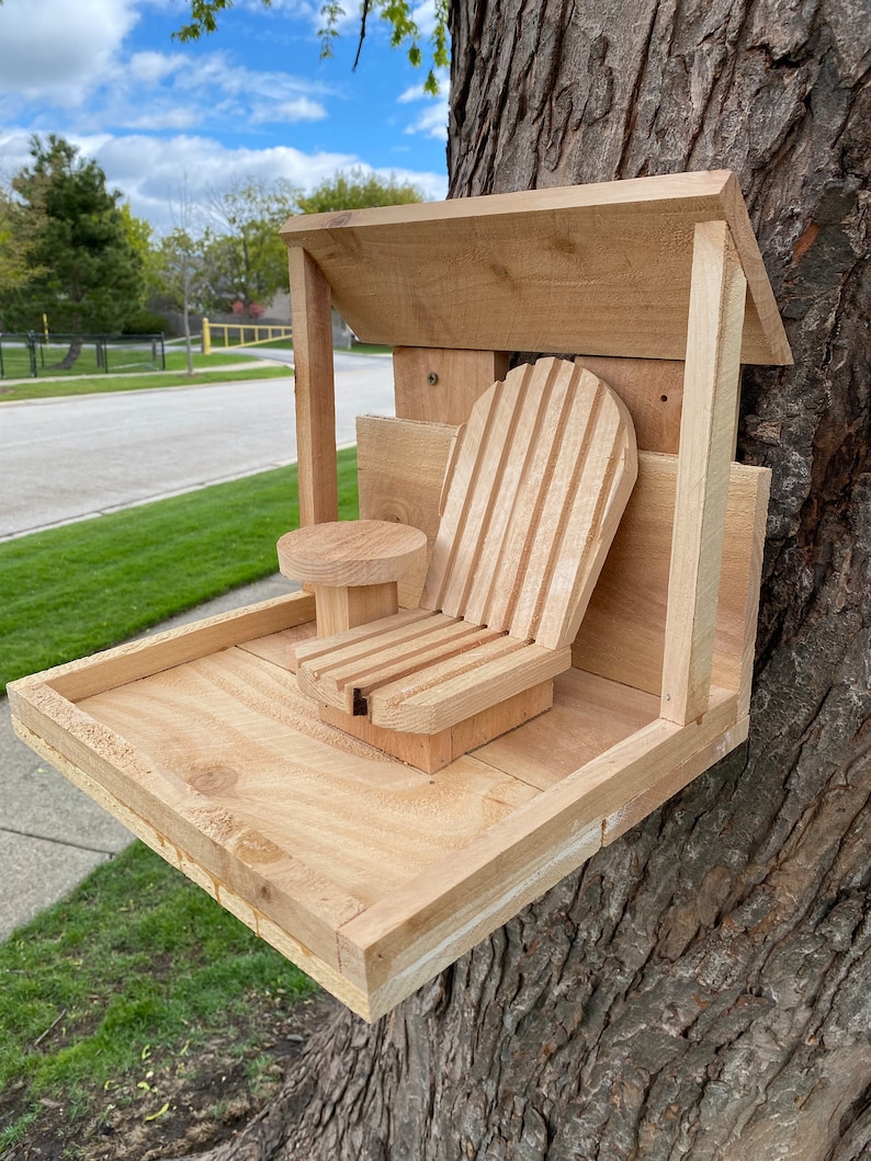 May include: A wooden bird feeder with a small chair attached to the front. The feeder has a roof and a tray for birdseed. The feeder is mounted to a tree trunk.