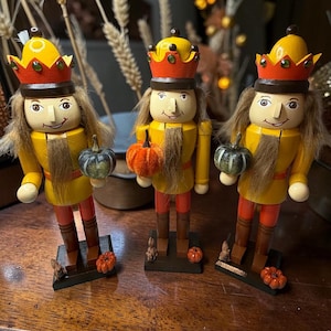 May include: Three wooden nutcracker figurines wearing yellow and orange crowns with green accents. Each figure has brown hair, a beard, and holds a small pumpkin. They stand on dark rectangular bases decorated with tiny orange pumpkins and squirrels.
