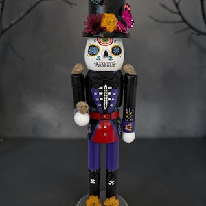 May include: A nutcracker figure painted as a skeleton with a white skull face, wearing a black top hat adorned with a blue butterfly, a pink butterfly, and flowers. The figure wears a black jacket with purple accents, a red belt, and purple pants.