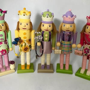 May include: Five wooden nutcracker dolls with painted faces, each wearing a crown and holding accessories. The dolls have different outfits, including floral aprons, skirts, and purses. The color palette includes pink, green, yellow, purple, and red.