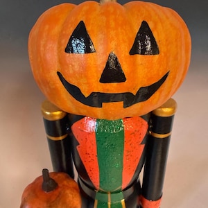 May include: A Halloween-themed nutcracker figurine with a pumpkin head and a small pumpkin accessory. The pumpkin head is orange with black facial features. The figurine's body is black with red, green, and gold accents.