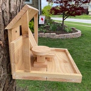 May include: A wooden bird feeder shaped like a house with a small deck and a miniature wooden chair and table. The feeder is attached to a tree trunk.
