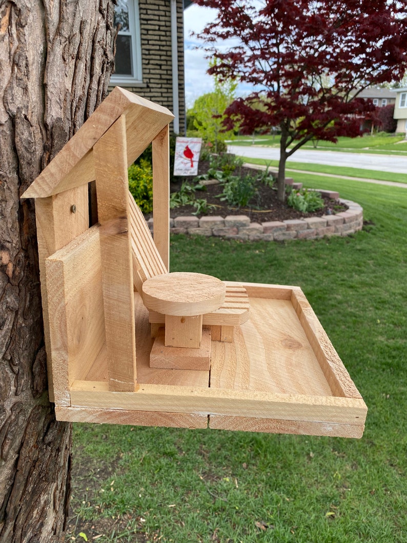 May include: A wooden bird feeder shaped like a house with a small deck and a miniature wooden chair and table. The feeder is attached to a tree trunk.