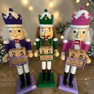 May include: Three wooden Christmas nutcrackers with white hair and smiling faces. Each figure wears a different colored crown and uniform: purple, green, and pink. Each holds a block calendar displaying "25 DAYS TIL CHRISTMAS".