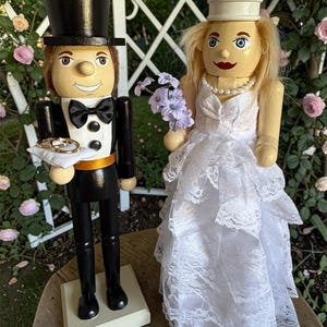 May include: Wooden bride and groom nutcrackers. The groom wears a black tuxedo with a top hat and holds a pillow with rings. The bride wears a white lace wedding dress and a crown, holding a bouquet of purple flowers. The figures are on a wooden surface.