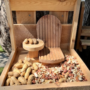 May include: A wooden bird feeder with a small wooden chair and table inside. The feeder is filled with peanuts and peanut shells.