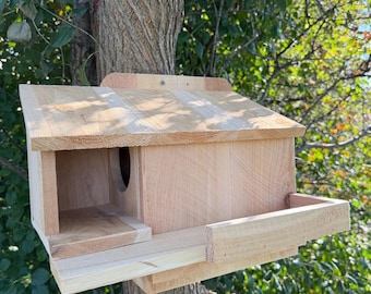 May include: A light-colored wooden birdhouse featuring a slanted roof, a rectangular entrance opening, and a flat front platform. The structure is constructed from natural wood planks with visible grain, designed for outdoor mounting.