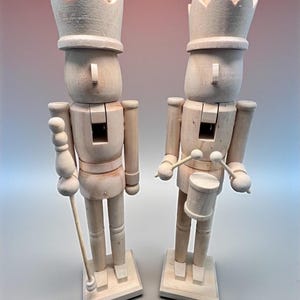 May include: Two unfinished wooden nutcrackers. One holds a staff, the other a drum and drumsticks. Both wear crowns and stand on square bases. The figures are light brown and have a classic design.