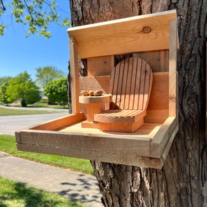 May include: A wooden bird feeder with a small chair and a tray for food. The feeder is attached to a tree trunk.
