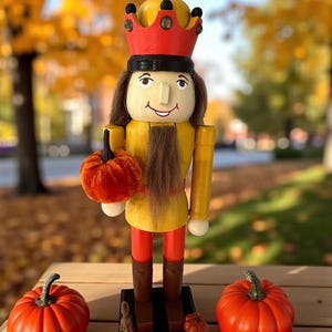 May include: A wooden nutcracker doll with a yellow and red crown, holding a small orange pumpkin. The doll has a yellow jacket, red pants, and brown boots. Two pumpkins sit on the table, with fall foliage in the background.