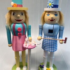 May include: Two wooden nutcrackers, each with blonde hair and a painted face. One wears a pink skirt and holds a cat on a scratching post. The other wears a blue and white checkered apron and holds a rake. Both have hats.