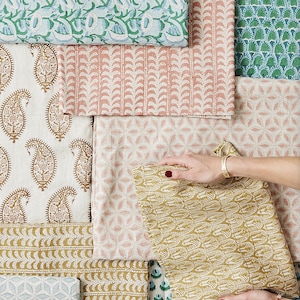 May include: An assortment of patterned fabrics in various colors and designs. The fabrics include floral, paisley, and geometric patterns in shades of green, blue, pink, and gold. A person's hands are holding a folded piece of gold fabric.