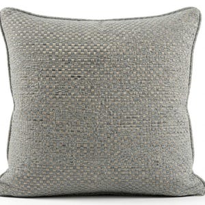 May include: A square throw cushion featuring a textured, woven fabric in shades of grey and beige. The cushion has a distinct piped edge detail around the perimeter, creating a structured border that frames the intricate, multi-tonal basketweave pattern.