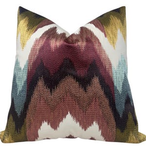 May include: Square cushion featuring a zig-zag pattern in burgundy, brown, blue, gold, and white. The cushion has a textured finish and a neutral background. The cushion is designed for decorative use.