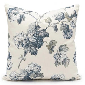 Könnte beinhalten: Quadratisches Zierkissen mit floralem Muster. Das Kissen hat einen cremefarbenen Hintergrund mit einem blau-grauen Blumendesign. Das Blumendesign umfasst Blätter und Blütentrauben.