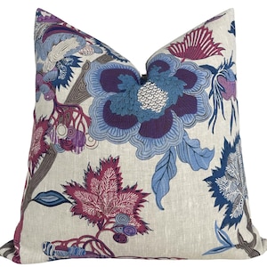 Puede incluir: Un cojín cuadrado de tela beige similar al lino con un estampado floral a gran escala en tonos morado oscuro, magenta y varios tonos de azul. El diseño incluye motivos botánicos intrincados y hojas detalladas.