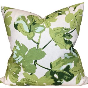 May include: A square throw pillow featuring a white background with a repeating pattern of large, stylized fig leaves in various shades of light green, medium green, and dark green. The fabric has a visible weave texture throughout the design.