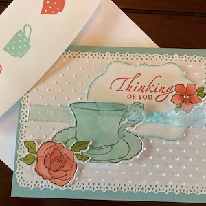 May include: A light blue card with a white lace border and a white background with a light blue teacup and saucer, a pink rose, and a white ribbon. The text "Thinking of You" is written in cursive on the card.