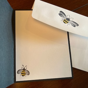 K&ouml;nnte beinhalten: Eine wei&szlig;e Karte mit schwarzem Rand und einer Bienen-Illustration auf der Vorderseite. Die Karte ist ge&ouml;ffnet, um einen leeren wei&szlig;en Innenraum zu zeigen. Ein wei&szlig;er Umschlag mit einer Bienen-Illustration ist enthalten.