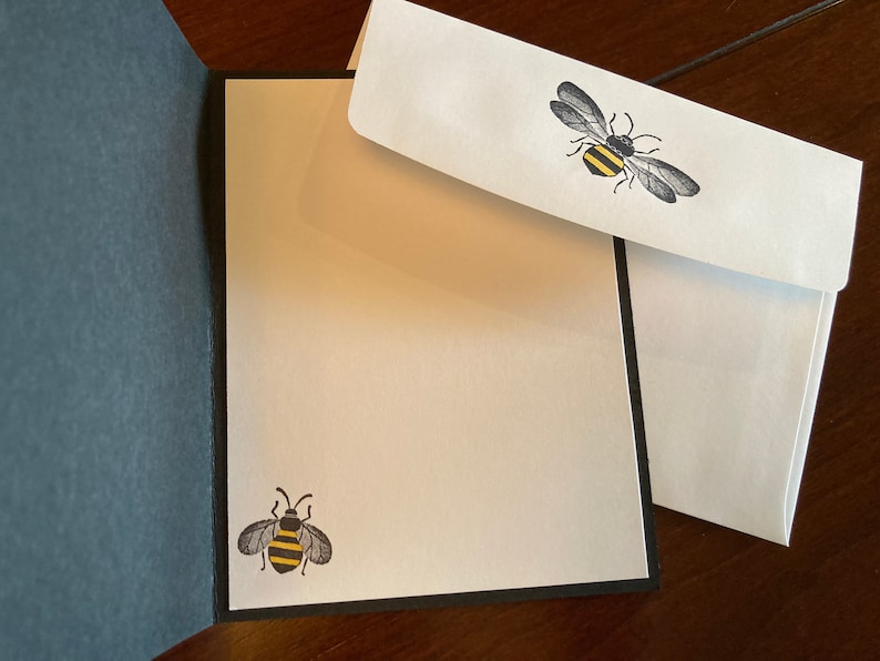 K&ouml;nnte beinhalten: Eine wei&szlig;e Karte mit schwarzem Rand und einer Bienen-Illustration auf der Vorderseite. Die Karte ist ge&ouml;ffnet, um einen leeren wei&szlig;en Innenraum zu zeigen. Ein wei&szlig;er Umschlag mit einer Bienen-Illustration ist enthalten.