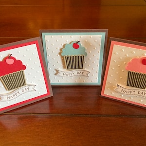 May include: Three handmade greeting cards featuring cupcake designs. Each card has a white background with a dotted pattern, a cupcake with a cherry on top, and a banner that reads "HAPPY DAY". The cupcakes are red, blue, and pink.