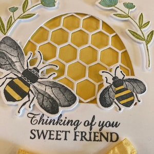 K&ouml;nnte beinhalten: Eine wei&szlig;e Karte mit einem gelben Wabenmuster, zwei schwarzen und gelben Bienen und dem Text "Thinking of you Sweet Friend". Ein gelbes Band ist unten an der Karte gebunden.