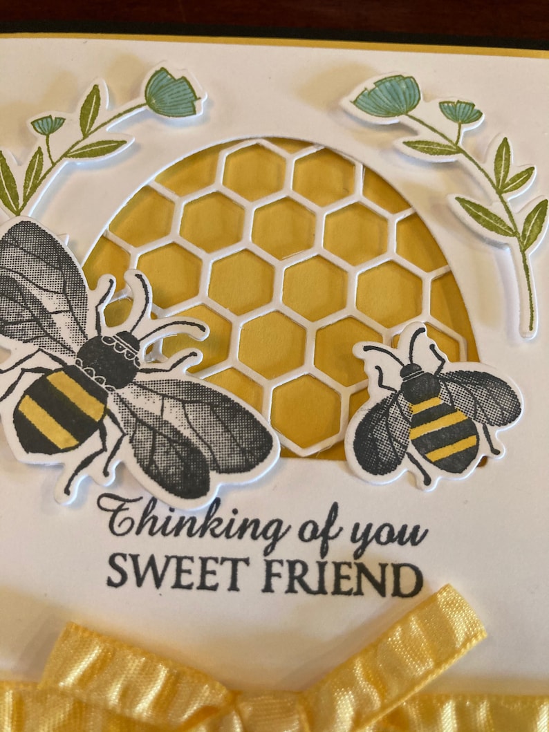 K&ouml;nnte beinhalten: Eine wei&szlig;e Karte mit einem gelben Wabenmuster, zwei schwarzen und gelben Bienen und dem Text "Thinking of you Sweet Friend". Ein gelbes Band ist unten an der Karte gebunden.